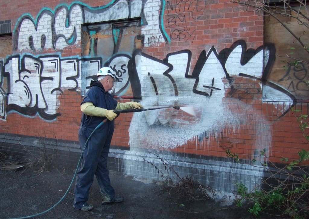 An older man wearing dark blue coveralls and a white hard hat is using a pressure washer wand to remove graffiti on a brick wall.